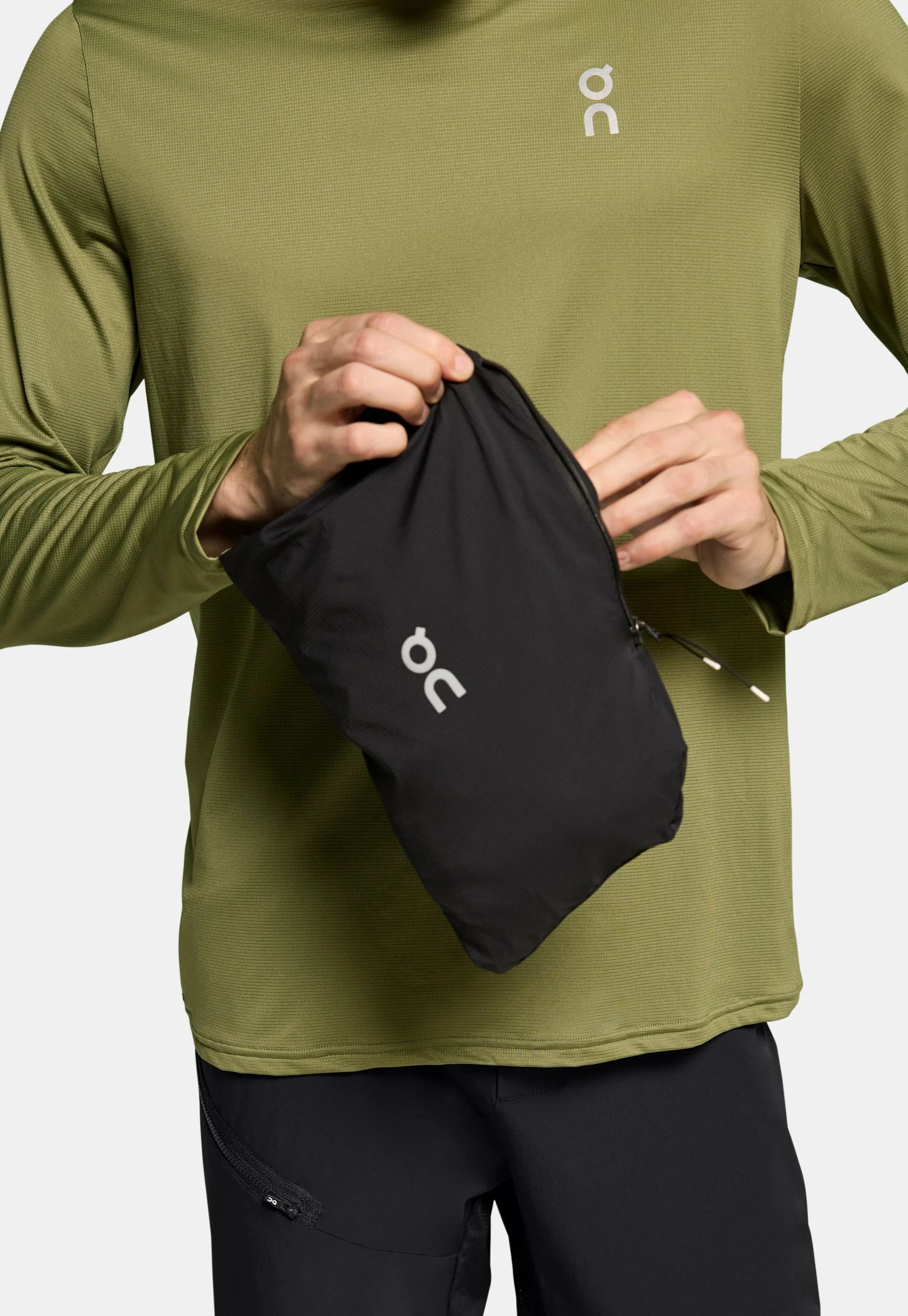 Una leggera pochette nera viene tenuta da una persona, con chiusura a zip e un logo bianco. L'individuo indossa una maglietta a maniche lunghe di un verde oliva strutturato.