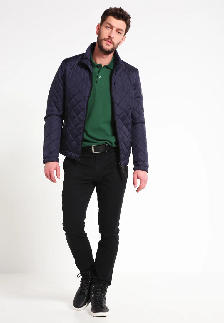 Giacca trapuntata blu navy con zip frontale, indossata sopra una polo verde. Abbinata a pantaloni neri e sneakers alte nere.