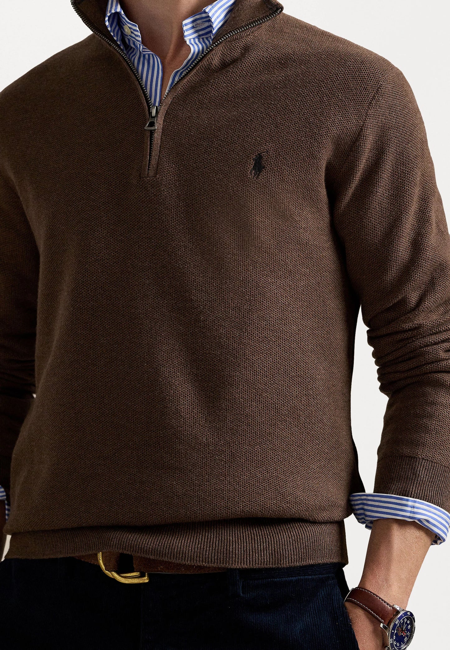 Suéter marrón con cuello de cremallera y tejido texturizado, que presenta un pequeño logo en el pecho, superpuesto a una camisa de rayas azul y blanca.