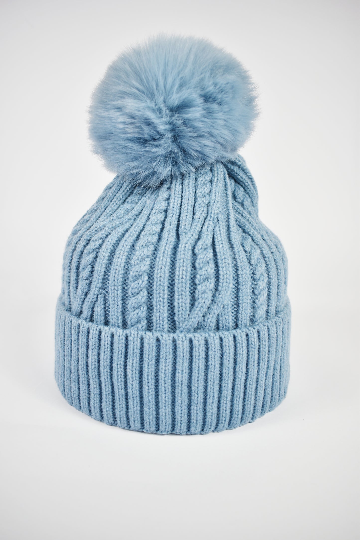 Berretto invernale da donna blu a trecce con risvolto e pompon in pelliccia ecologica su sfondo bianco.