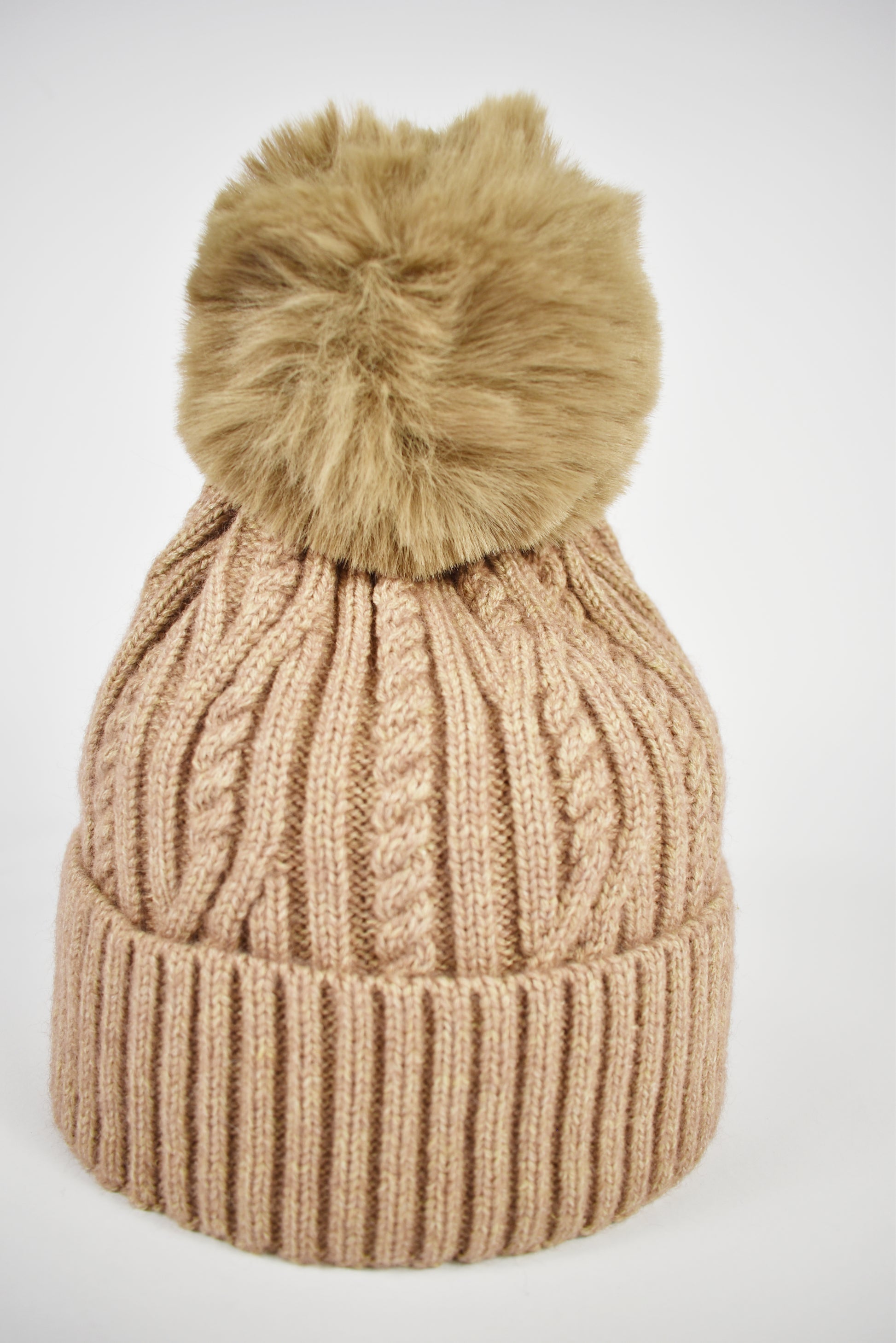 Berretto donna beige lavorato a trecce con risvolto morbido e pompon in pelliccia ecologica su sfondo bianco.