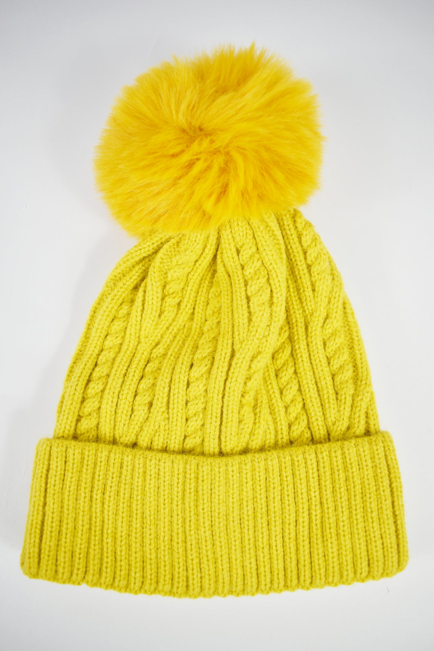 Cappello invernale giallo donna a trecce con risvolto e pompon in pelliccia ecologica morbida su sfondo bianco.