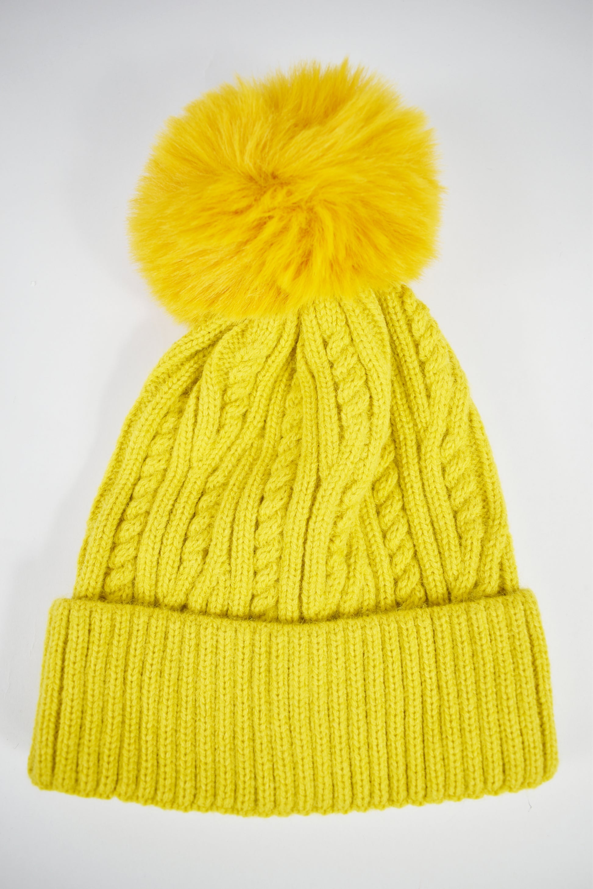 Cappello invernale giallo donna a trecce con risvolto e pompon in pelliccia ecologica morbida su sfondo bianco.