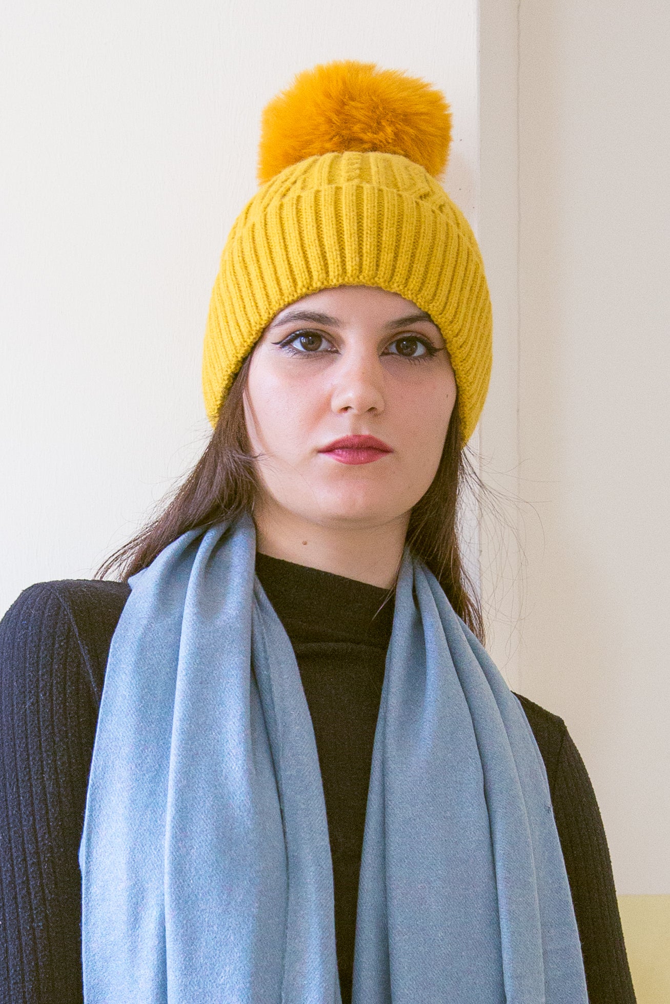 Donna indossa un berretto invernale senape a trecce con risvolto e pompon giallo in pelliccia ecologica.